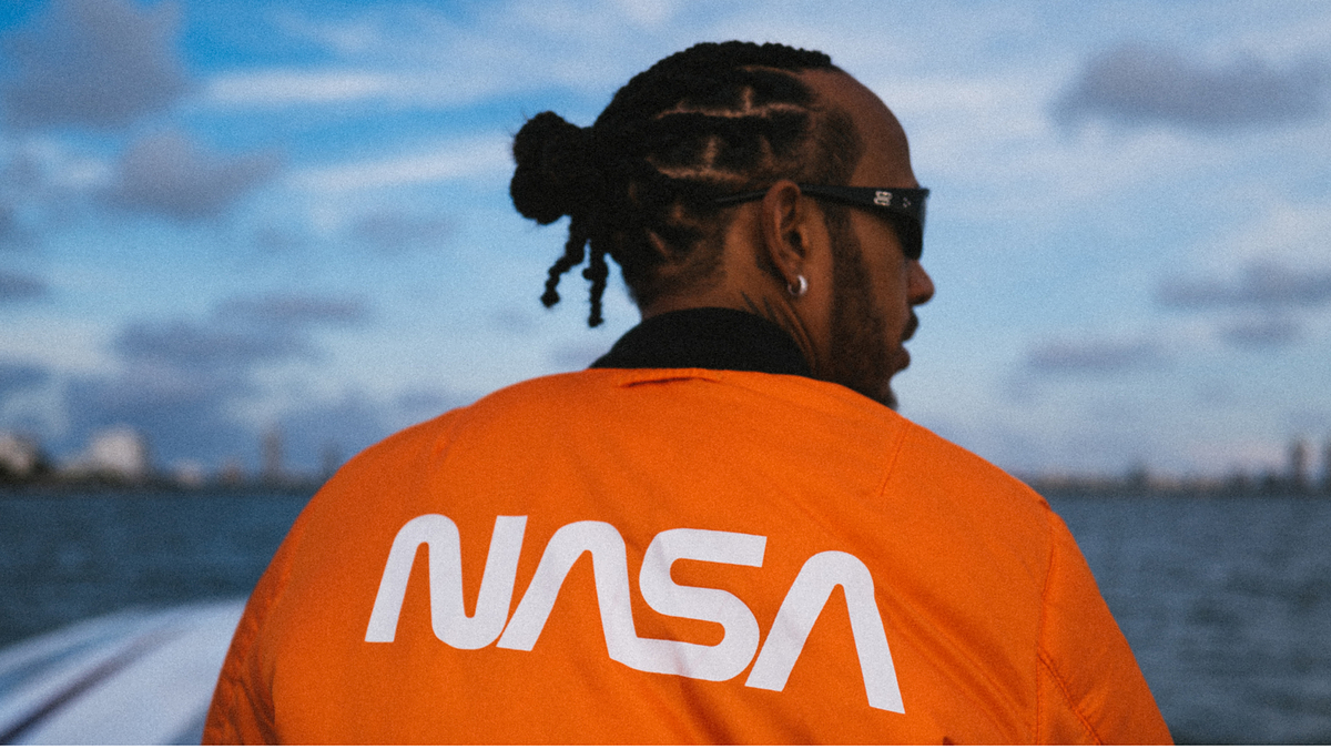 Person in orange NASA jacket facing away, looking at scenic sky with water in the background