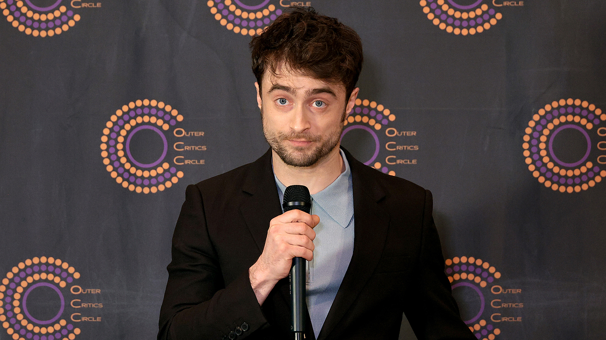 Man in a suit at a press event, holding a microphone, backdrop with logos