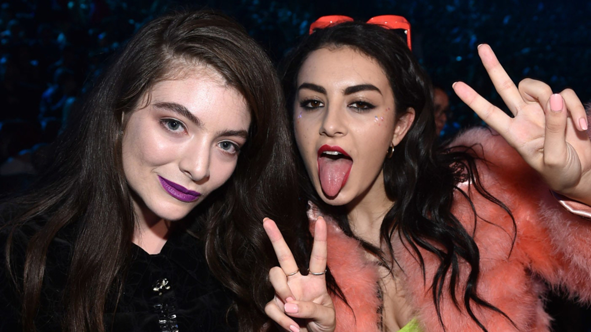 Lorde and Charli XCX make peace signs at a music event. Charli wears a fur jacket and sunglasses, while Lorde is in a dark top