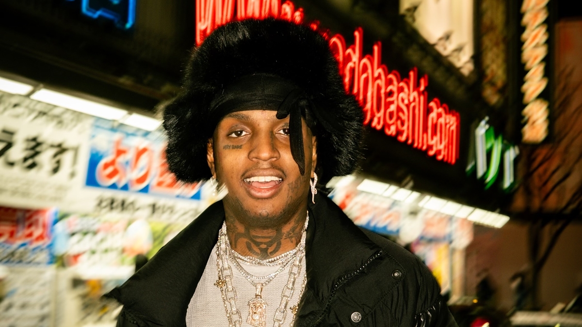 Rapper Polo G stands outdoors, smiling, wearing a black puffer jacket, layered necklaces, and a furry hat, with brightly lit signs in the background
