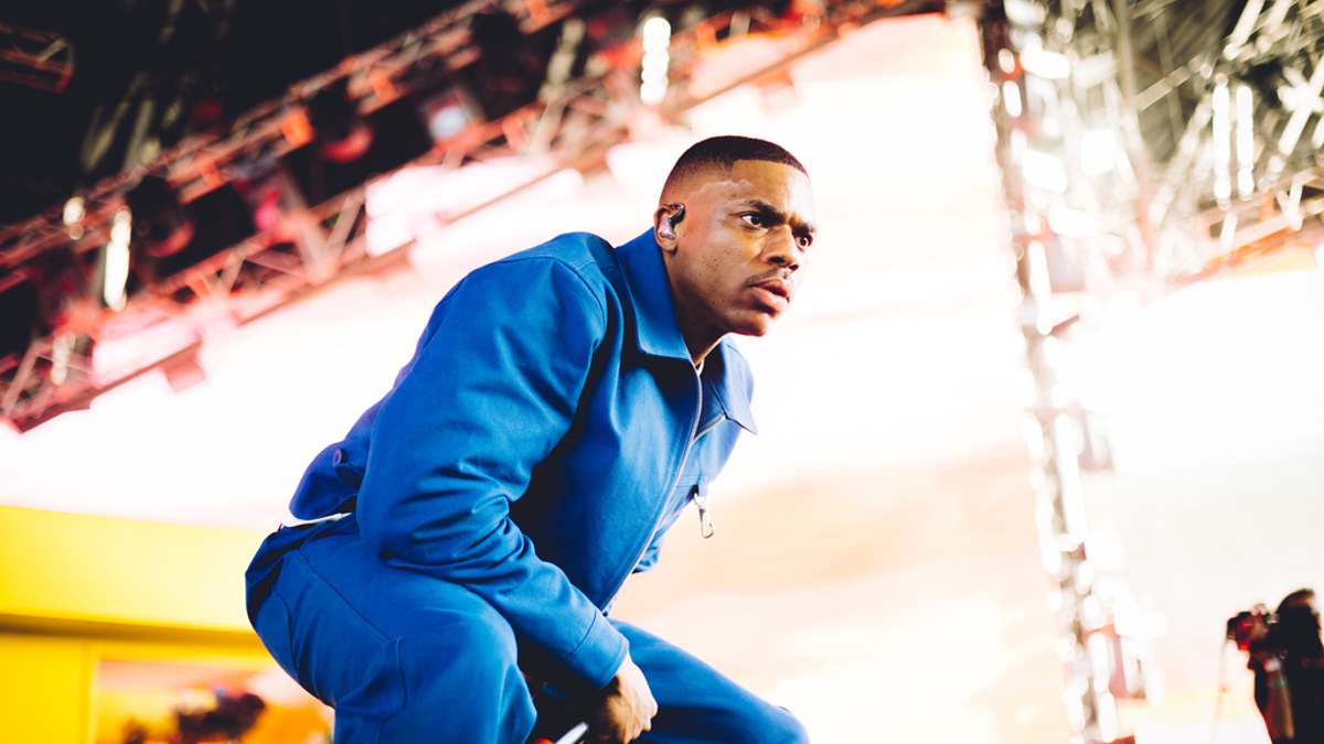 Vince Staples performing on stage, wearing a jumpsuit and holding a microphone. Bright lights and concert equipment are visible in the background