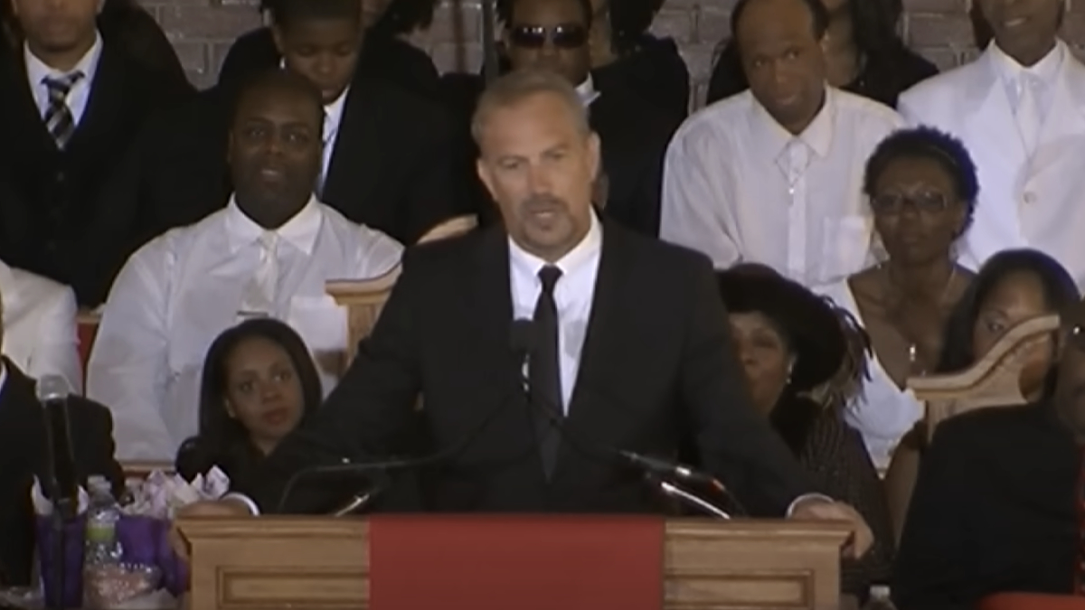 Kevin Costner speaks at a podium during a formal event, surrounded by attendees dressed in formal attire