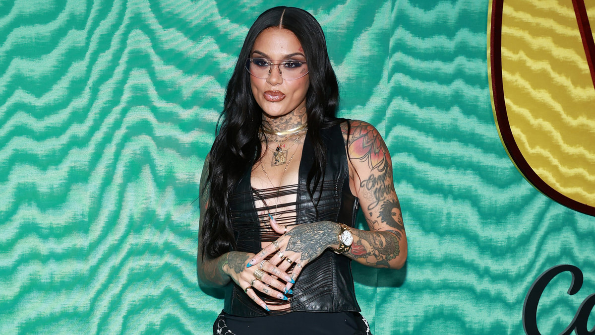 Kehlani in a black strappy top and black pants with visible tattoos on her arms and hands, posing on a patterned backdrop at a music event