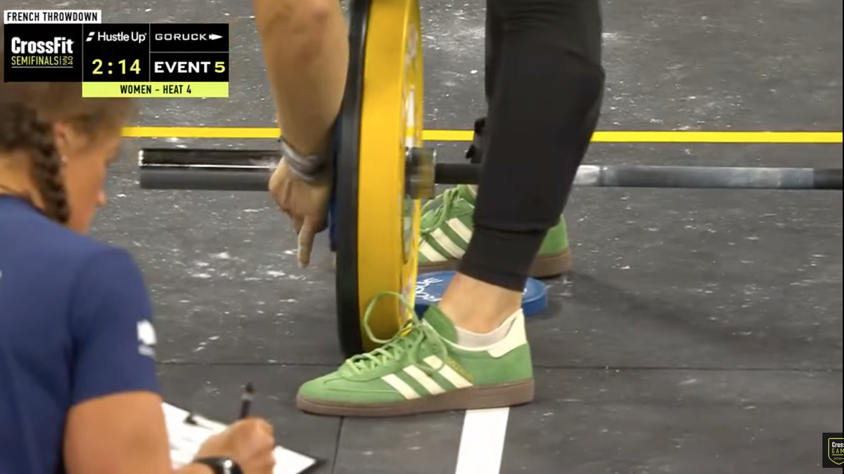 Close-up of a weightlifter adjusting weights on a barbell at the French Throwdown CrossFit Semifinals Event 5. Timer shows 2:14. An official is visible writing on a clipboard
