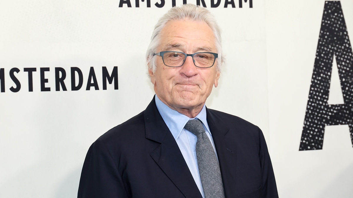 Robert De Niro smiles wearing glasses, a dark suit, light blue shirt, and tie, at the "Amsterdam" event backdrop
