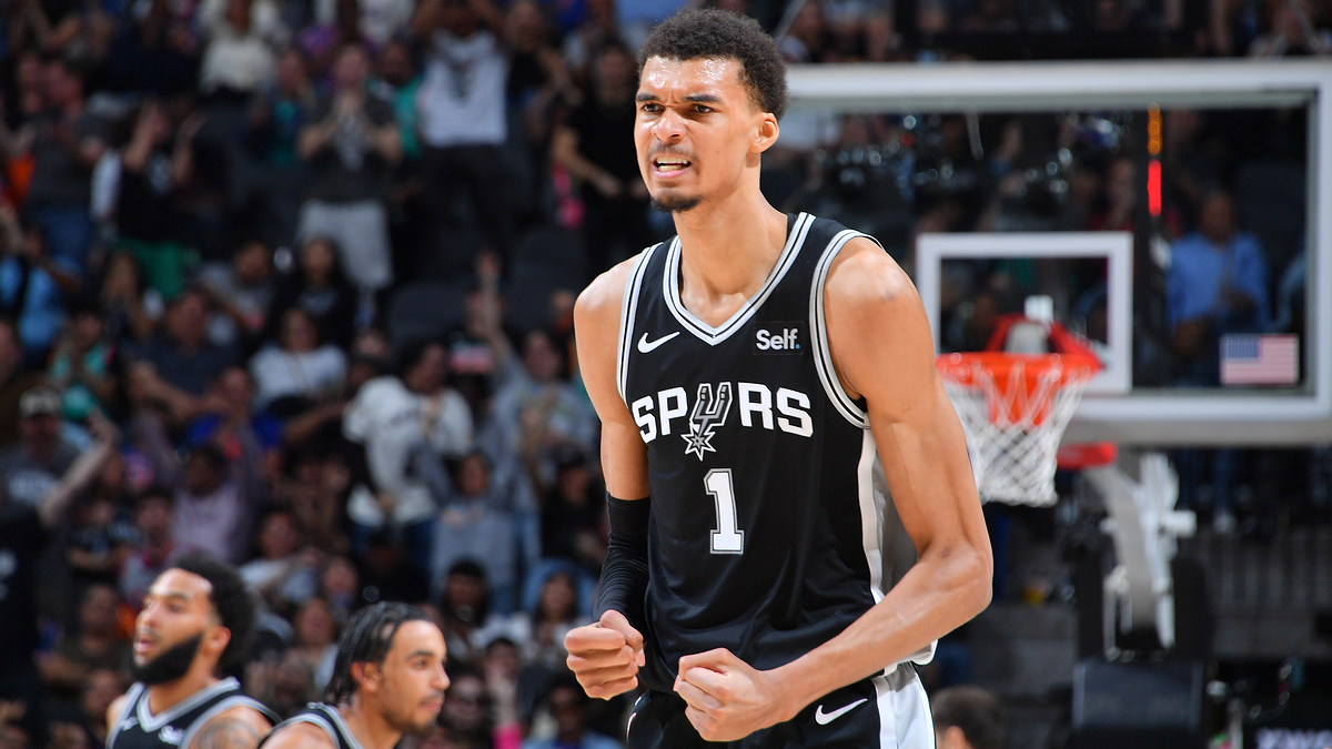 A basketball player in a Spurs uniform mid-game, on the court with focused expression, other players in the background