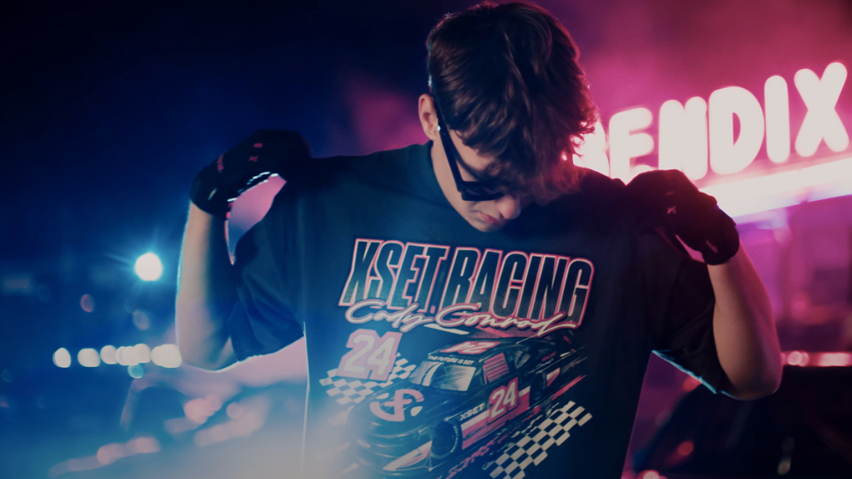 Person showing off a "KSET Racing Cup Series" T-shirt with a race car graphic. The person is wearing sunglasses and gloves, with neon signs in the background