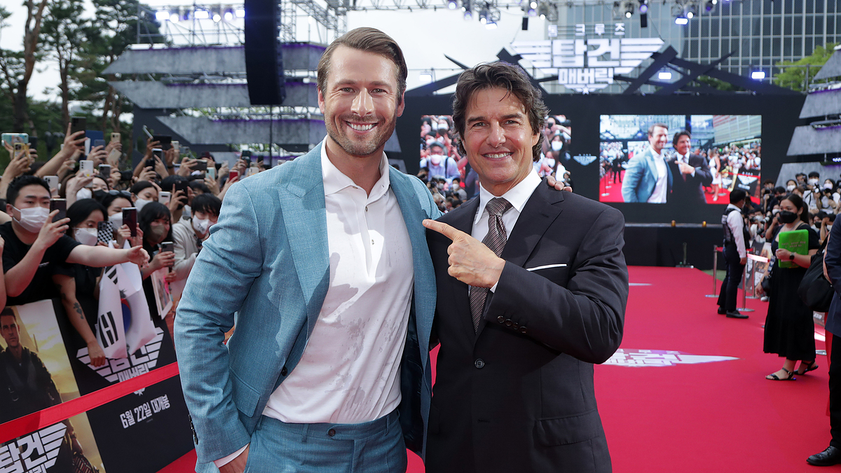 Glen Powell and Tom Cruise on a red carpet at a "Top Gun: Maverick" event, posing for photos with fans in the background. Glen Powell wears a blue suit and Tom Cruise a black suit
