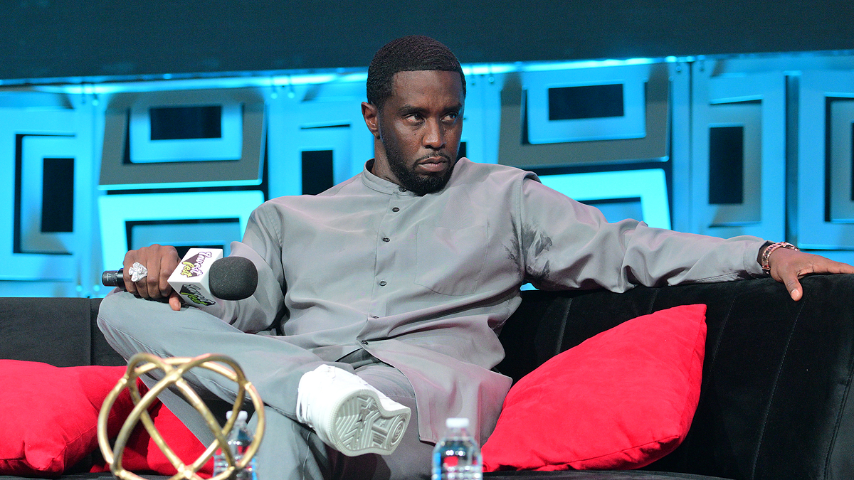 Sean "Diddy" Combs sits on a black couch with red pillows, wearing a light-colored suit. He holds a microphone in one hand and looks intently forward