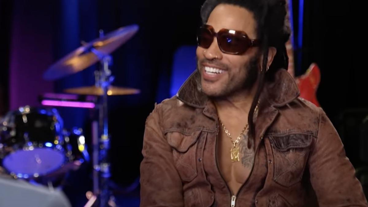 Lenny Kravitz, wearing sunglasses and a brown leather jacket, smiles during an interview in a studio with a drum set and guitar in the background