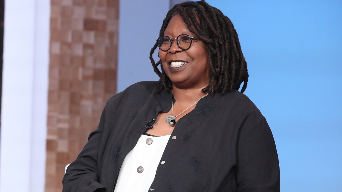 Whoopi Goldberg smiles in a black jacket and layered necklaces on a talk show set