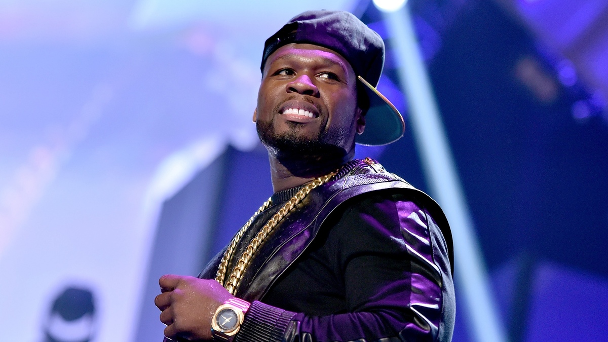 50 Cent on stage wearing a black leather-sleeved jacket, multiple gold chains, a wristwatch, and a backward baseball cap. He is smiling