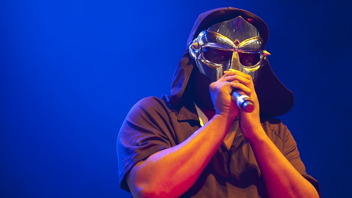 A performer, MF DOOM, is on stage wearing a metallic mask and holding a microphone. He is dressed in a dark-colored shirt with a hood