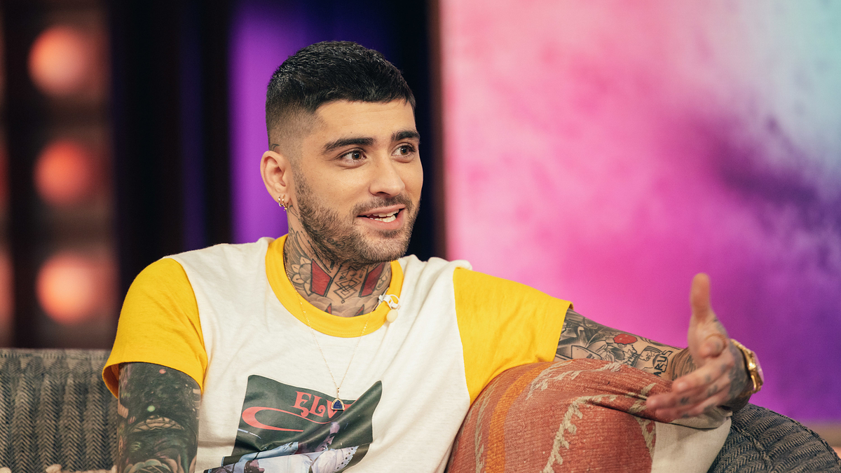 Zayn Malik sitting on a couch, animated while in conversation, wearing a graphic t-shirt and tattoos visible