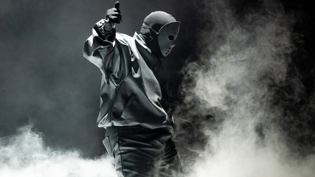 Music artist in mask and hoodie on stage with dramatic lighting and smoke, pointing upwards