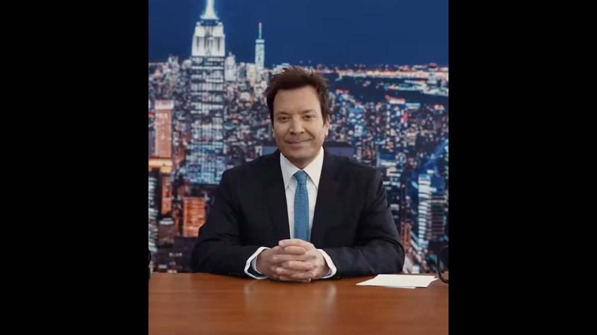 Jimmy Fallon sits at a desk in front of a cityscape background, smiling and wearing a black suit and tie