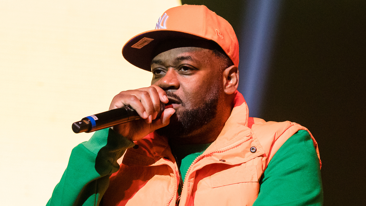 Ghostface Killah performing, wearing a puffy vest, cap, and green shirt while holding a microphone