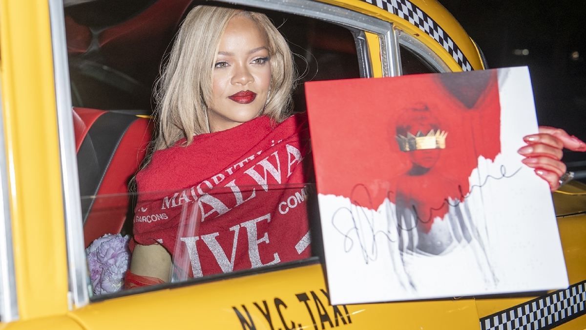 Rihanna in a taxi, holding a signed artwork featuring a red lipstick print of her lips. She wears a red scarf with intricate details