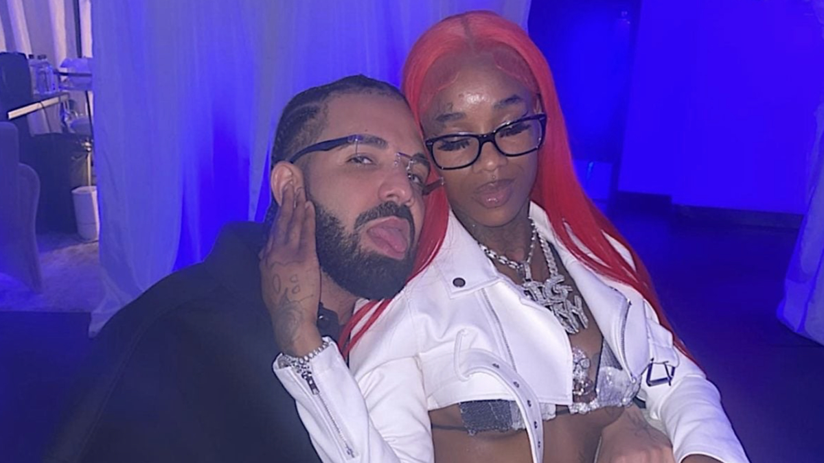 Drake and Sexyy Red pose together, smiling. Sexyy Red wears a silver and white outfit with a white jacket and glasses, both adorned with jewelry