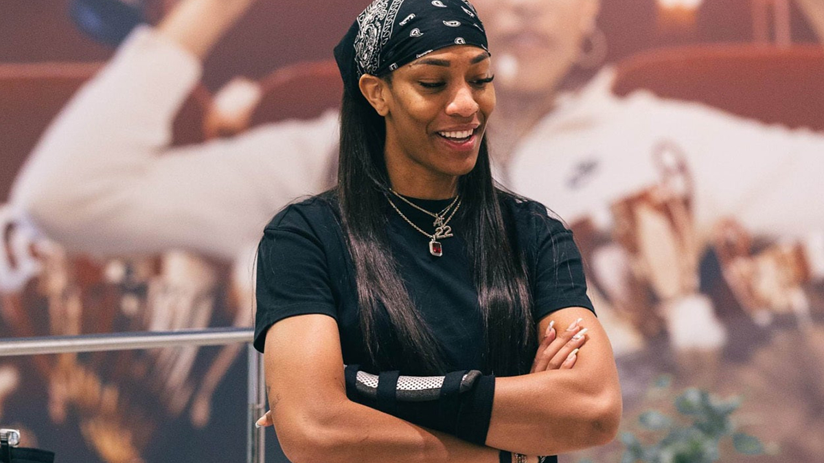 Person standing with crossed arms, wearing a headband, sleeveless shirt, and bracelets, with a poster in the background