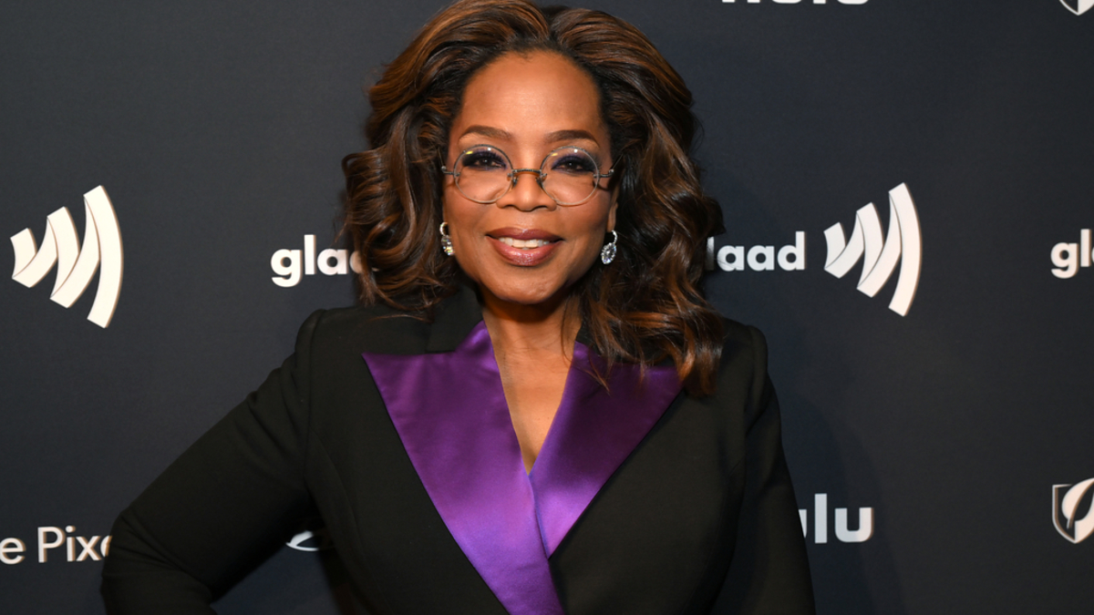 Oprah Winfrey posing with hands on hips, wearing a black suit with a purple collar detail, at a media event