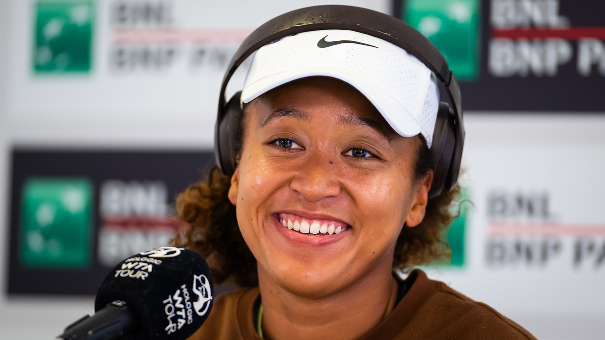 Naomi Osaka wearing a hat and smiling during a press conference