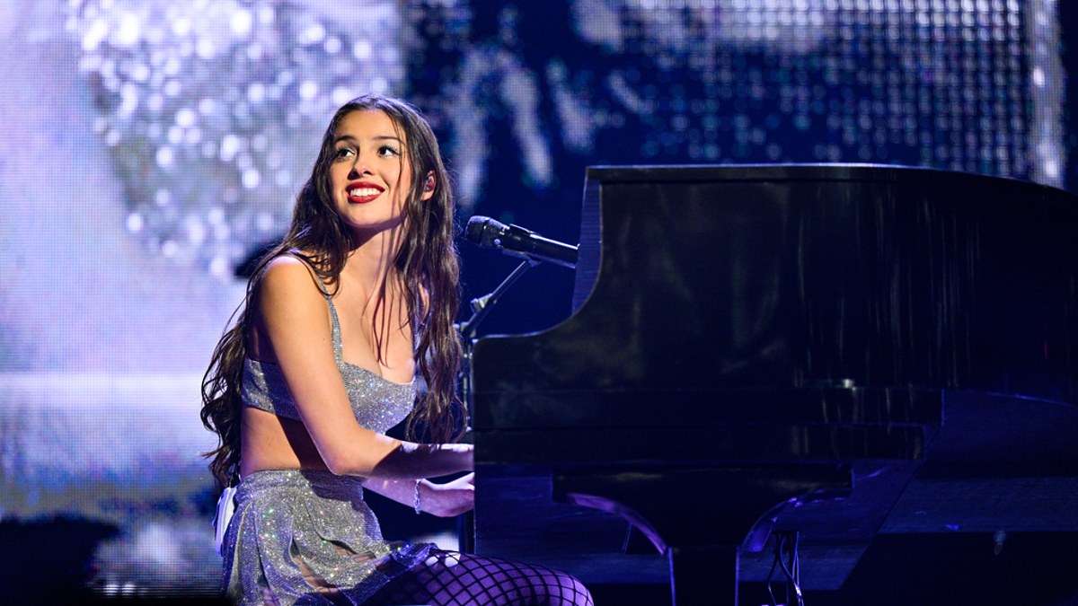 Performer at piano on stage with microphone, wearing a sparkling dress. Emotion through musical expression