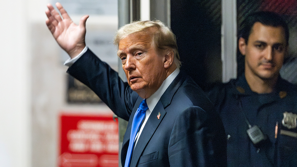 Donald Trump waves while standing next to a security officer. He is dressed in a suit and tie