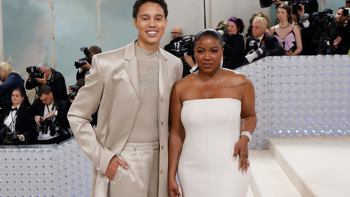 Two individuals posing at an event, one in a beige suit, the other in a white strapless dress. Photographers in the background