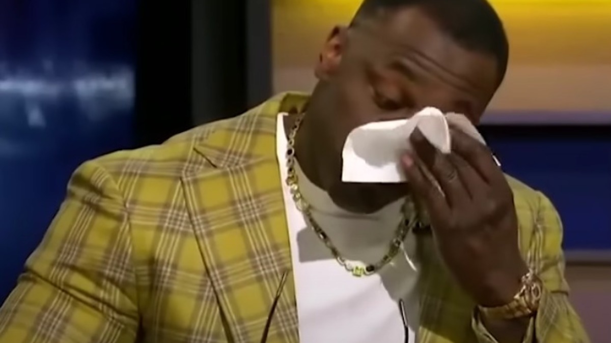 Shannon Sharpe wipes a tear while holding glasses, wearing a plaid jacket and white shirt, during a sports talk show