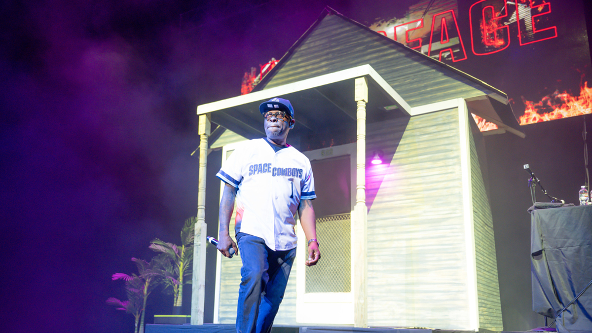 Performer in casual attire stands on stage with a house prop and "PEACE" sign in the background