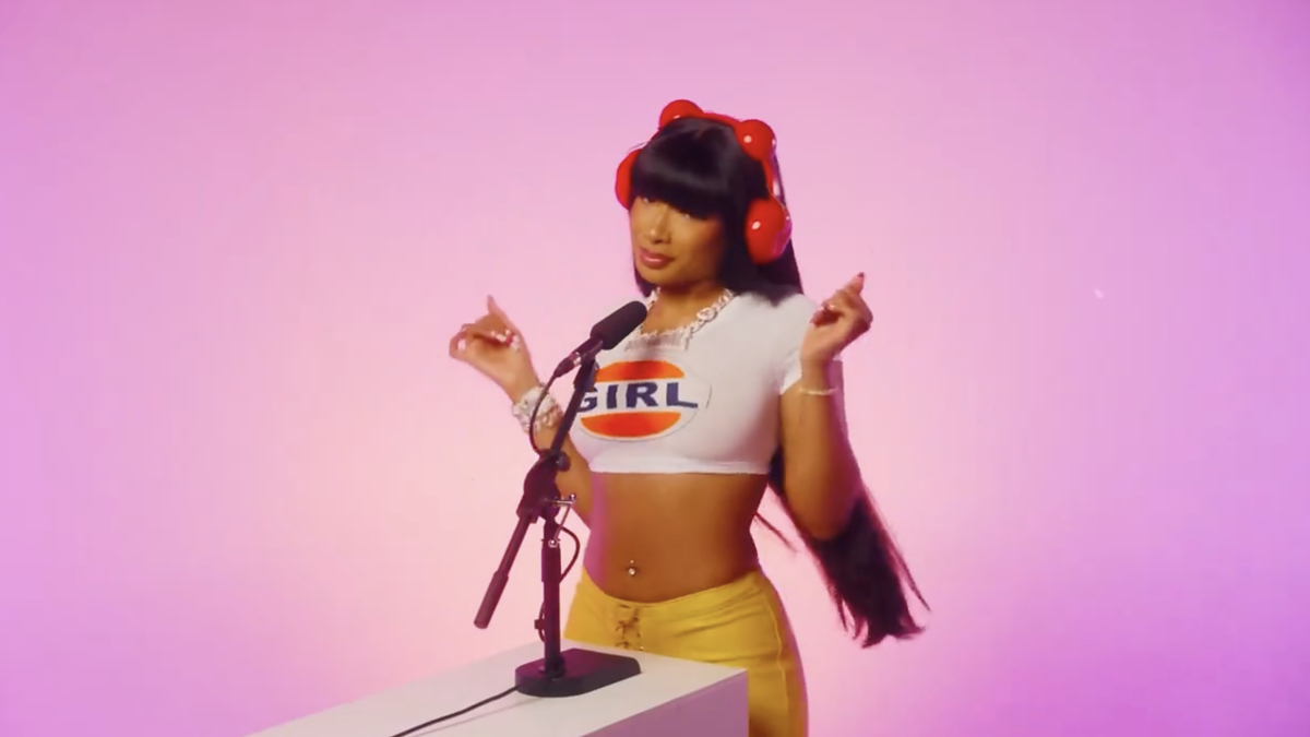Person on pink backdrop by mic wearing a cropped top with 'GIRL' print, yellow pants, and red hair accessories