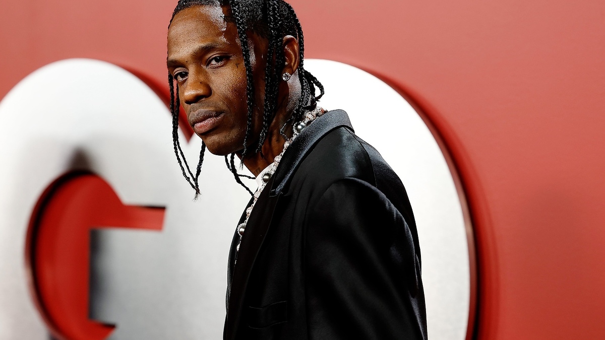 Travis Scott in a black suit with white shirt, standing in front of a large letter "G."