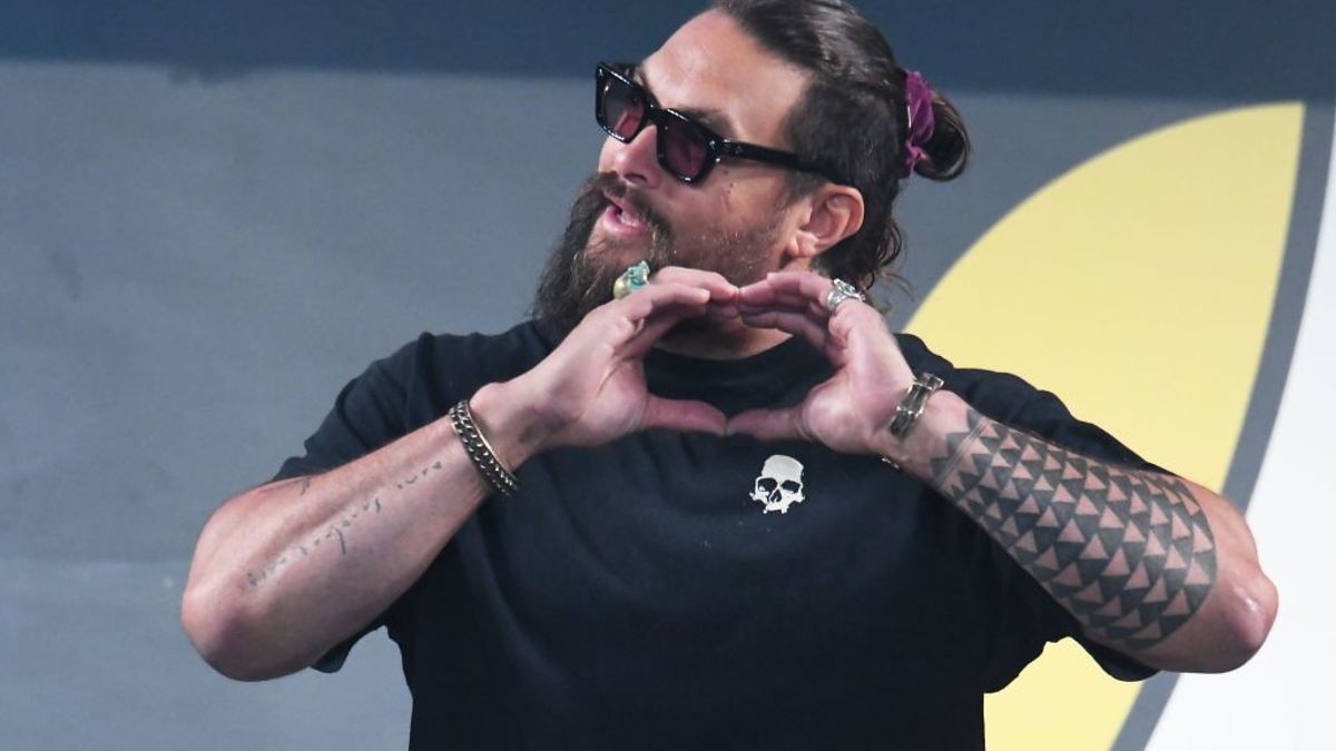 Jason Momoa makes a heart shape with his hands, wearing a black shirt, sunglasses, and a patterned arm tattoo, during a public appearance