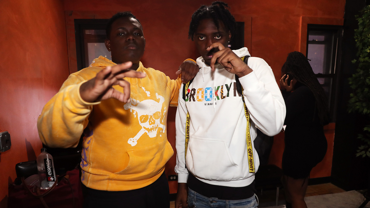 Two individuals pose together indoors. The person on the left wears a yellow hoodie with a white skull design, and the person on the right wears a white hoodie with "BROOKLYN" written on it