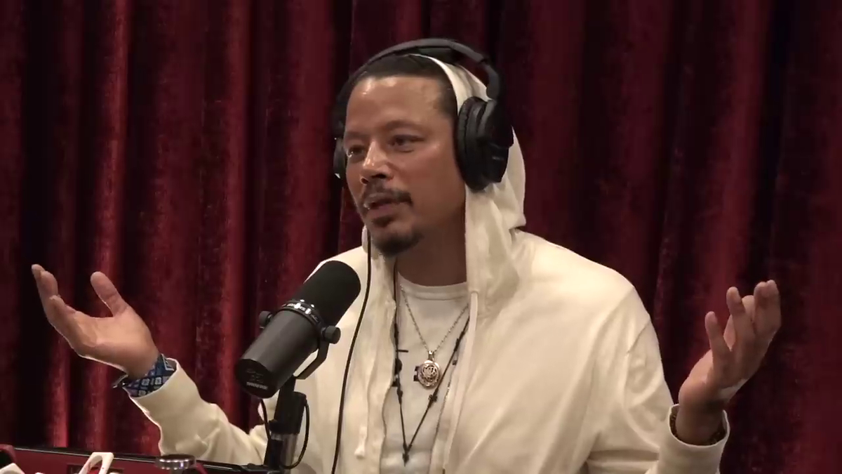 Person in a white outfit and headphones speaks into a microphone, gesturing with one hand. Red curtain in the background