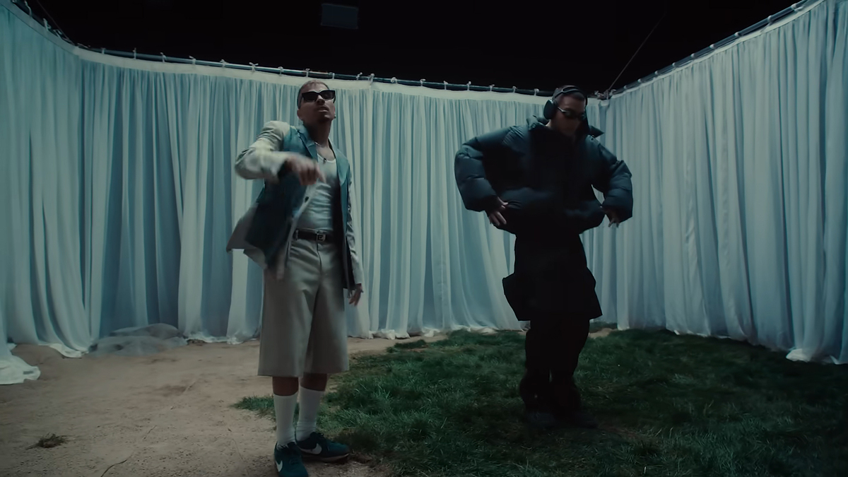 Two people dancing in a draped room, one in a long coat and sunglasses, the other in a blazer