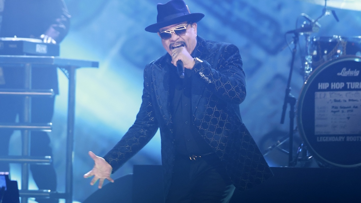 Singer performing on stage wearing a hat and patterned jacket, with a drummer in the background
