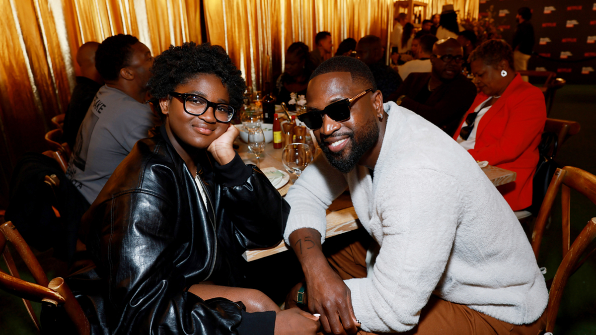 Dwyane Wade and Zaya Wade sit at a table at an event, dressed in casual chic attire. Other attendees are visible in the warmly lit background