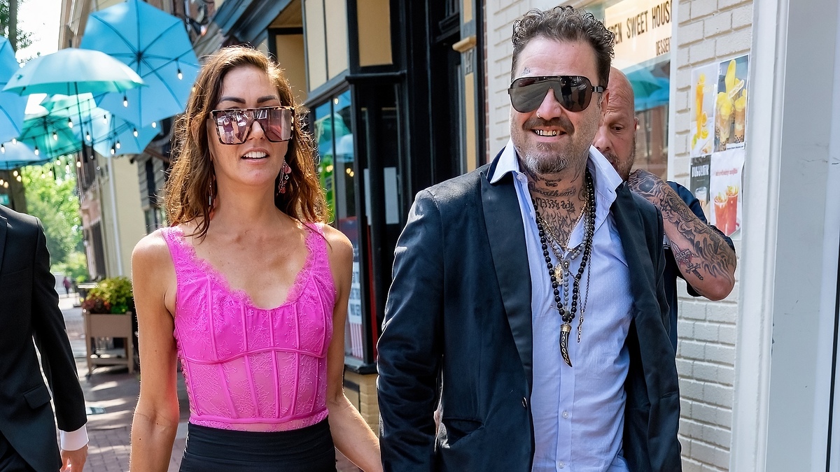 A man with tattoos and wearing sunglasses, a dark coat, and a white shirt walks beside a woman in a sleeveless top and sunglasses. Both are smiling