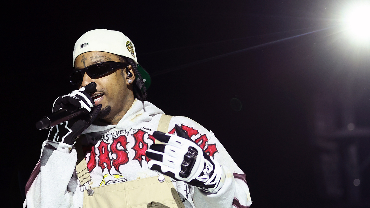 Music artist performs onstage, wearing a graphic tee, dungarees, and a cap, holding a microphone