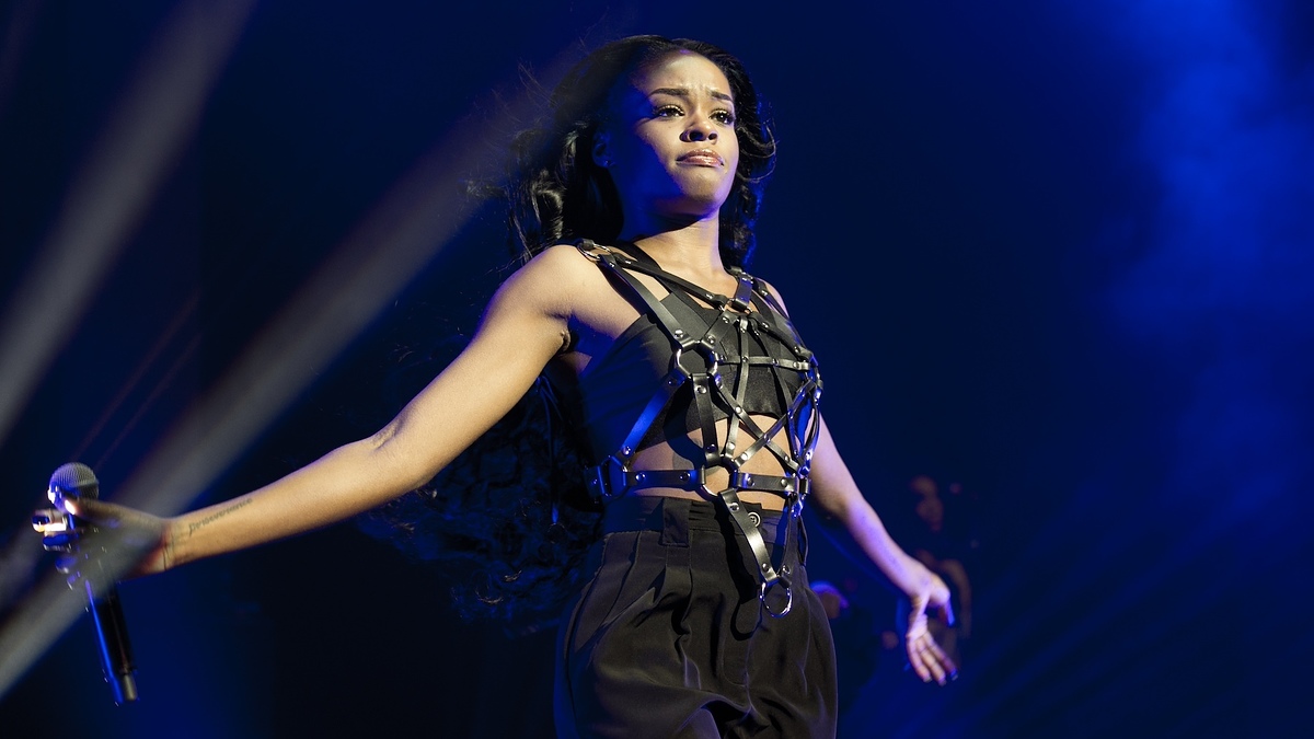 Azealia Banks performs on stage wearing a sleeveless top with a strappy harness and high-waisted pants. She has long hair flowing behind her