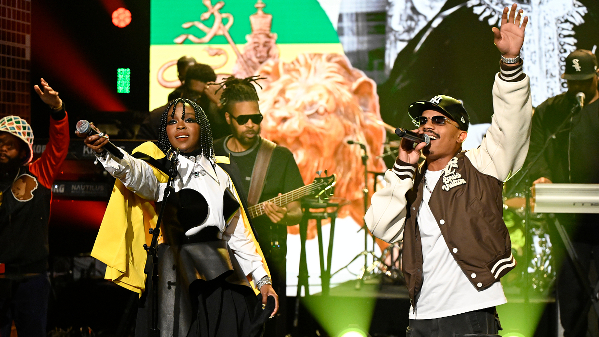 Two musicians perform on stage with a band, one in a black cap and jacket, the other with braids and a yellow top