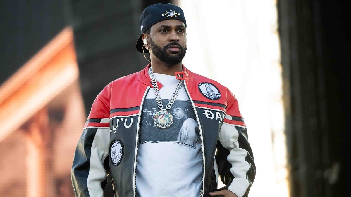 Man in a baseball cap and letterman jacket with graphic prints, wearing headphones around neck