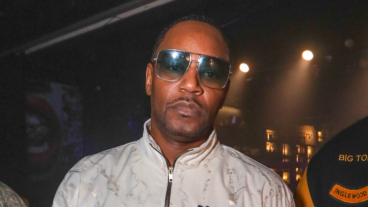 A man in a white patterned zip-up jacket and large sunglasses looks at the camera in a dimly lit environment