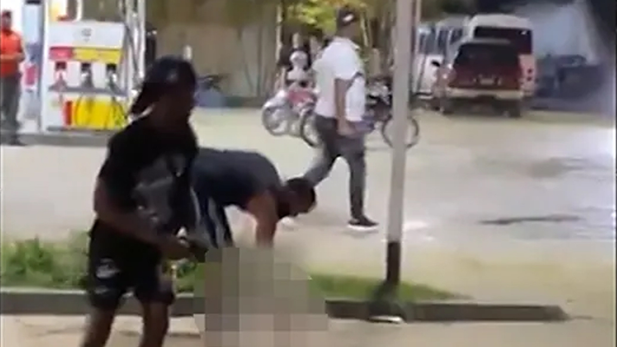Two individuals in a street altercation, with one person swinging at another. Background shows bystanders and vehicles