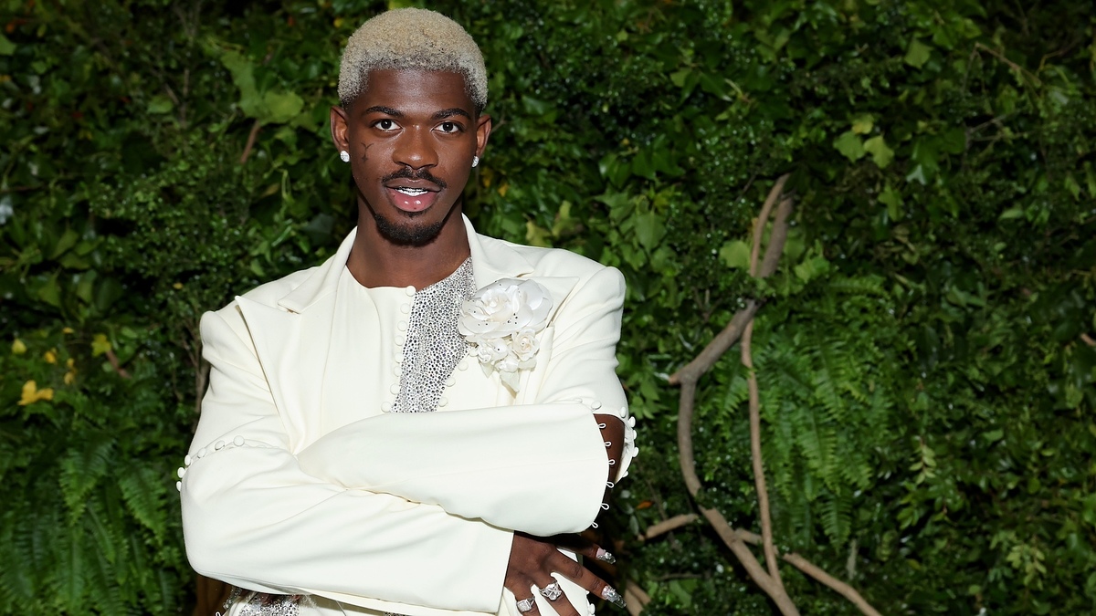 Lil Nas X poses outdoors wearing a cream-colored suit with a detailed floral brooch and sparkling glam jewelry. He stands against a backdrop of lush greenery