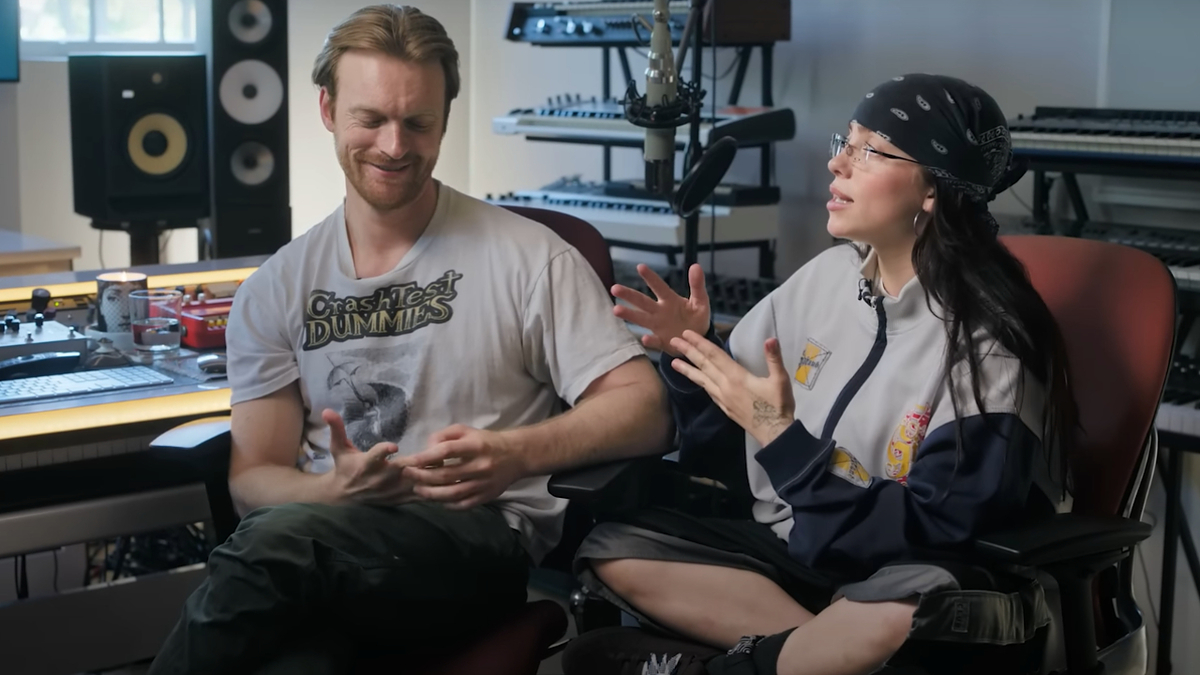 Finneas and Billie Eilish sit in a music studio, conversing animatedly. Finneas wears a casual T-shirt and pants. Billie wears a bandana, large jacket, and shorts