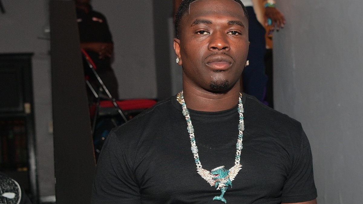 Man in a black shirt with a large bird-shaped necklace stands looking at the camera