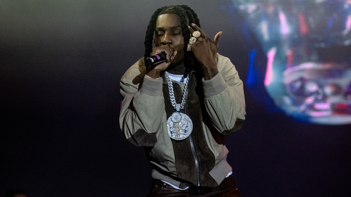 Rapper on stage performing, wearing heavy chain necklaces and rings, holding a microphone
